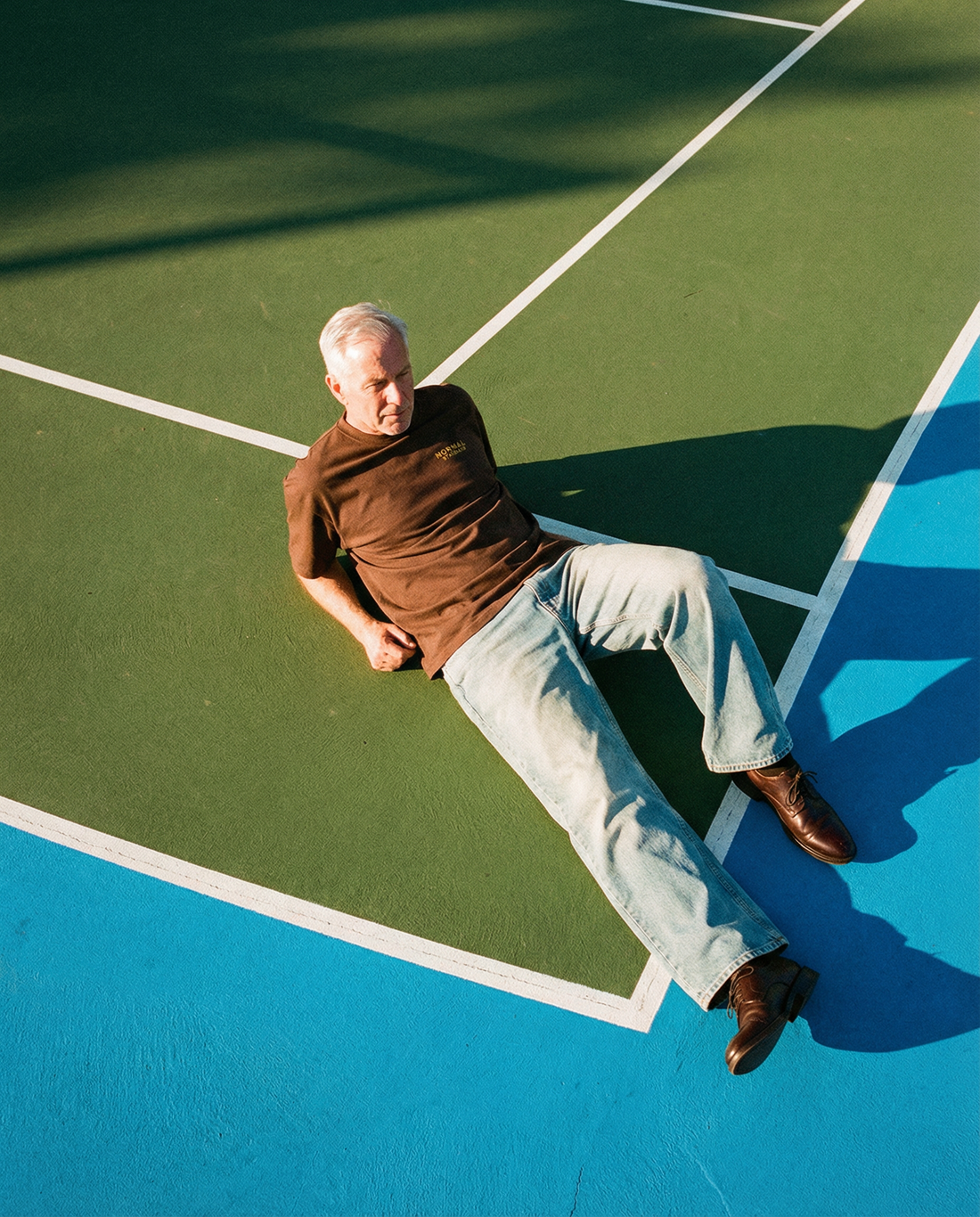Man sitting on a colorful outdoor tennis court with geometric white lines and long shadows in warm sunlight wearing Normal Standard Club Oversize t-shirt.
