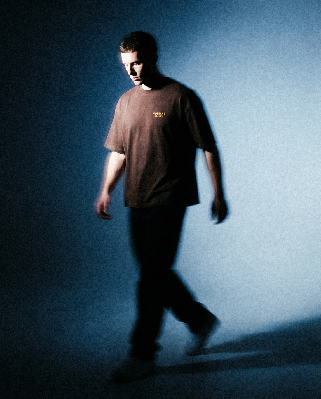 A young man in a brown oversized T-shirt walks across a dimly lit blue background, with soft motion blur and dramatic lighting casting shadows around him.