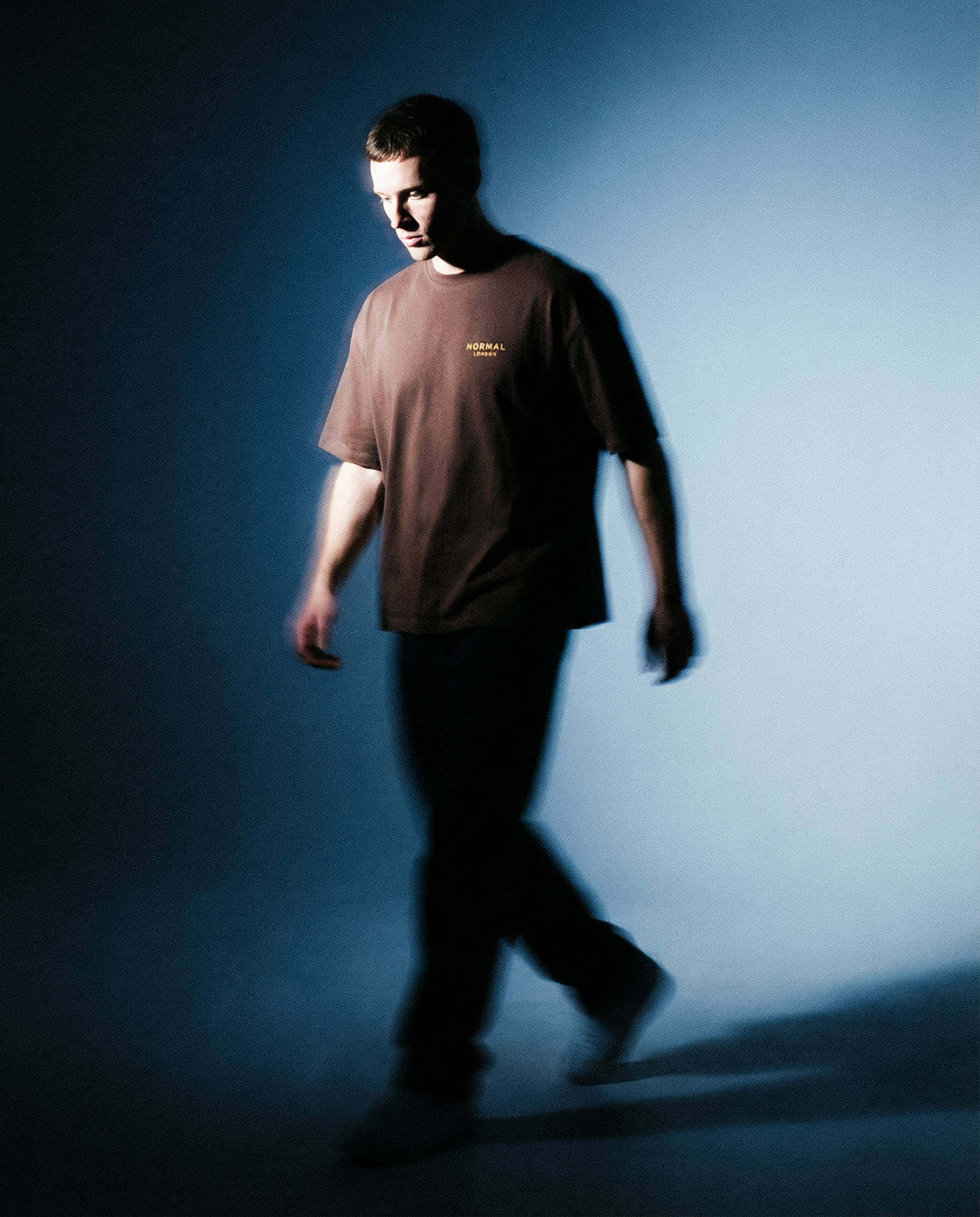 A young man in a brown oversized T-shirt walks across a dimly lit blue background, with soft motion blur and dramatic lighting casting shadows around him.