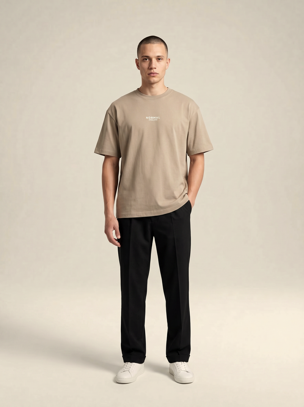 Man wearing a Brenton beige t-shirt and black pants on a plain background with the wording Normal England at the center of the chest.