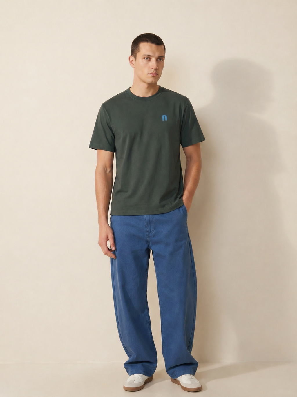 Man wearing a green t-shirt and blue pants against a beige background