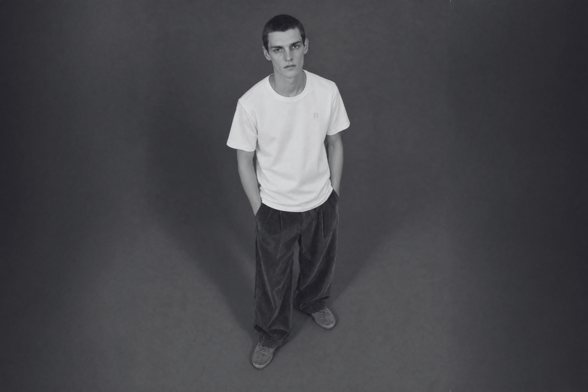 A man wearing a white crew-neck t-shirt and dark corduroy trousers, standing against a dark gray studio background.