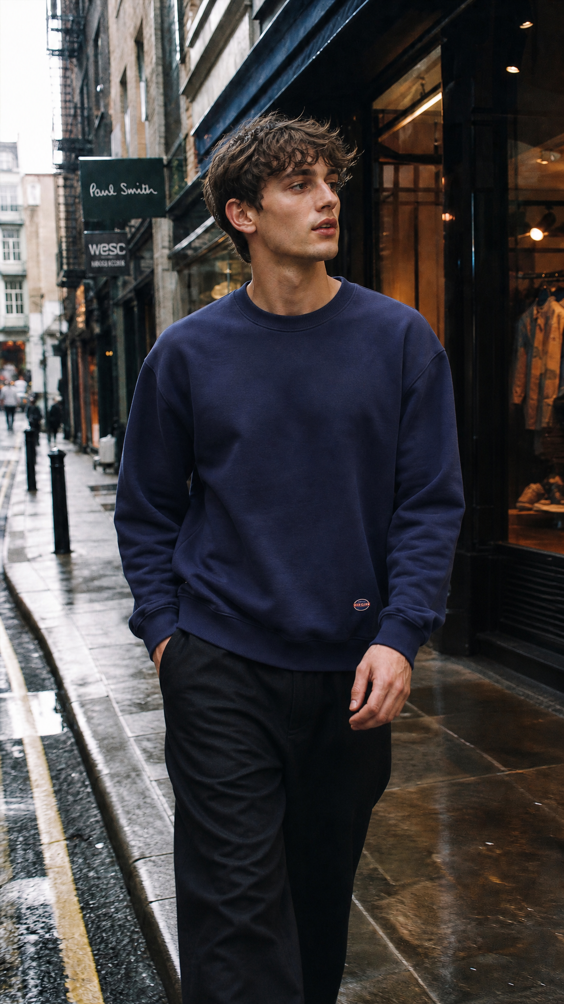 A man walking on a wet city street wearing a Normal Standard navy blue crewneck sweatshirt and black trousers, with storefronts in the background.