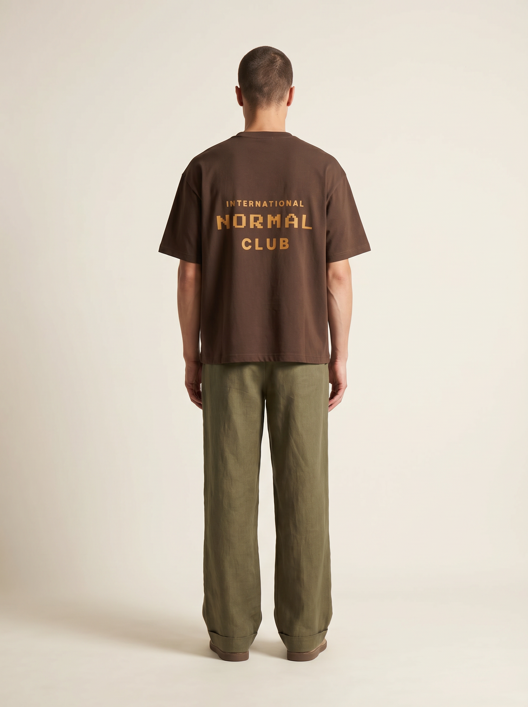 Dark Brkown  | Person wearing a brown t-shirt with 'International Normal Club' text on the back, standing against a plain background.  | Back model