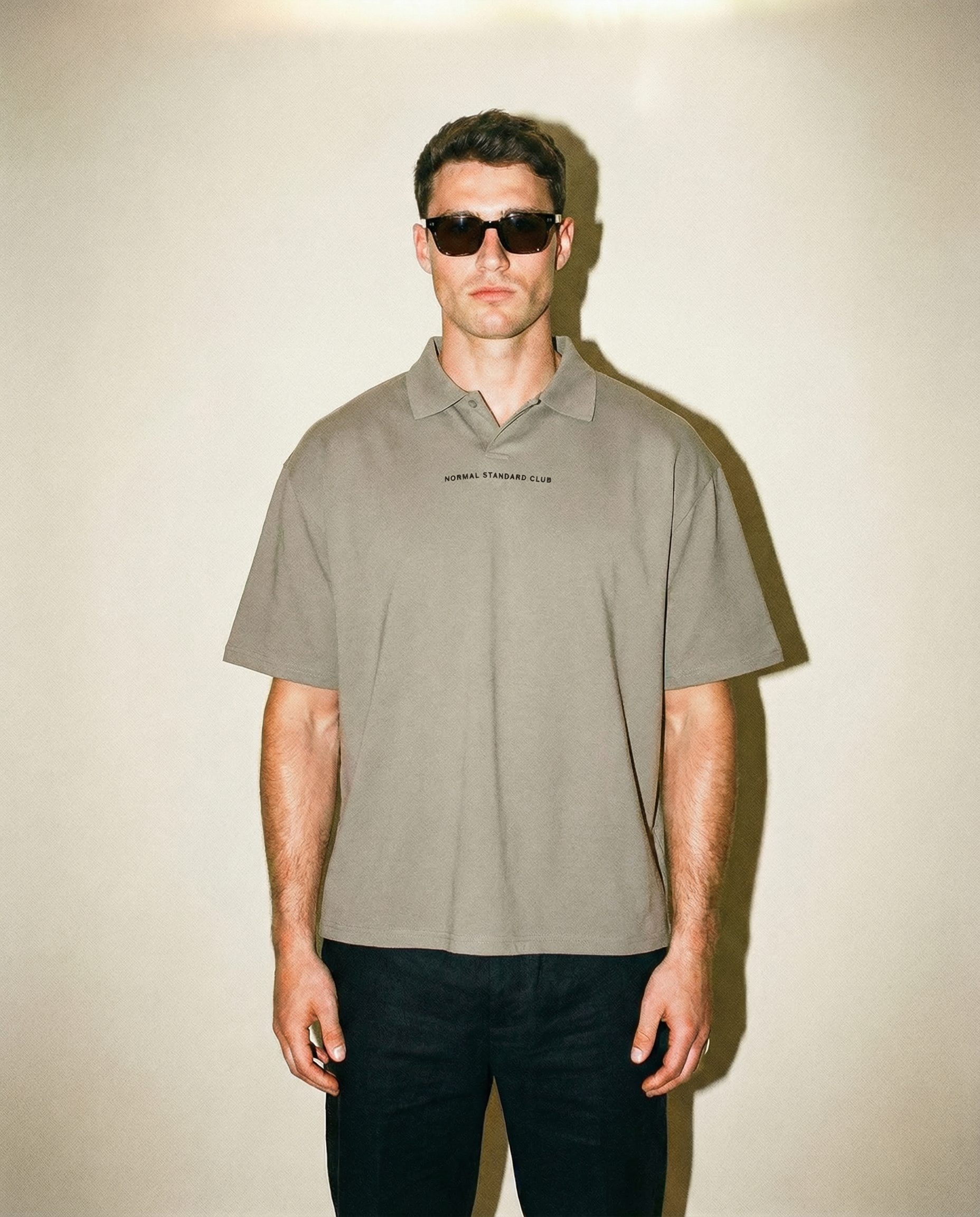 Man wearing a gray polo shirt with Normal Standard Club logo across the chest and dark sunglasses against a plain background