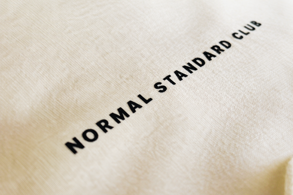 Texture text 'NORMAL STANDARD CLUB' on a beige tank-top.